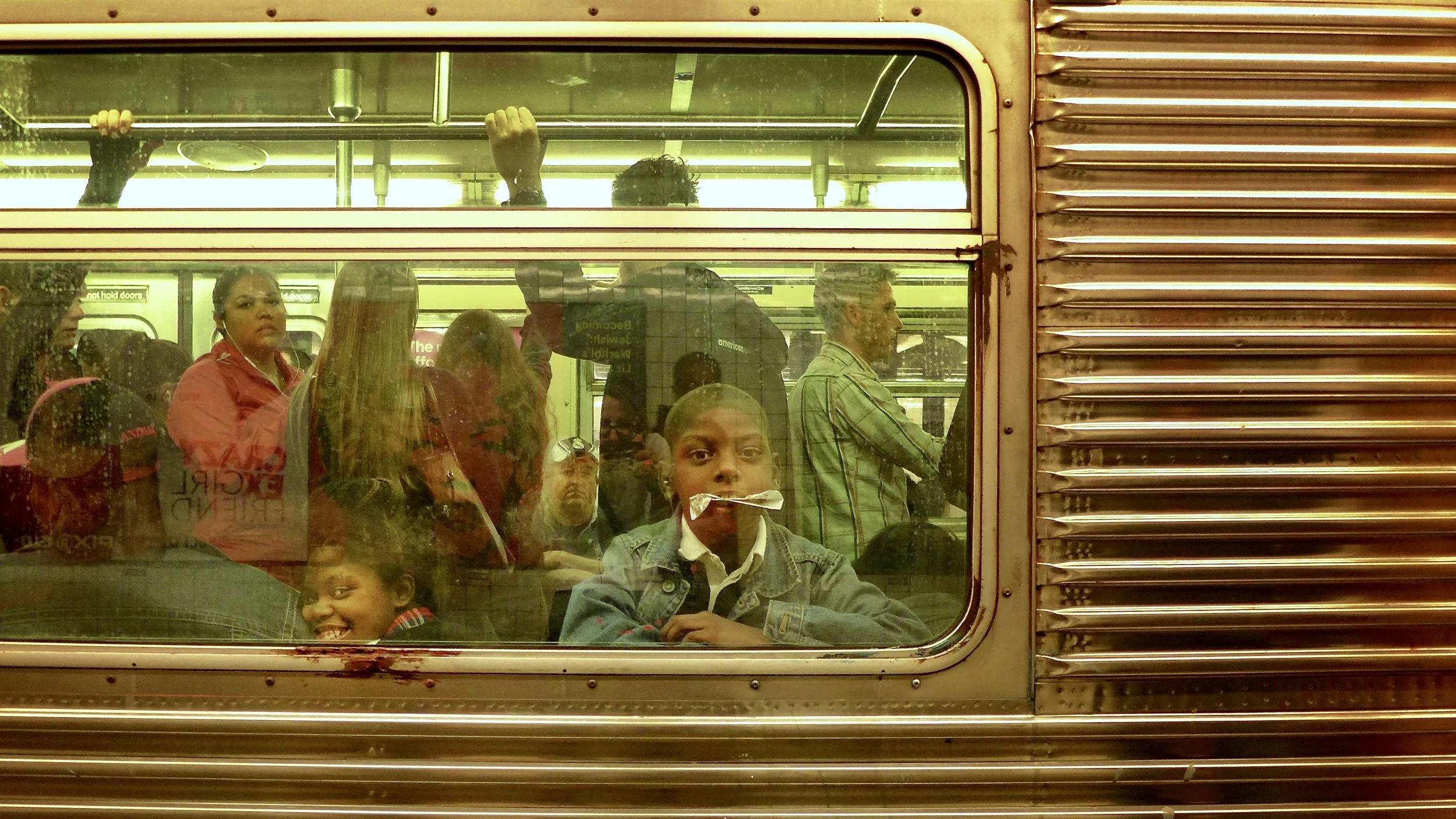 People sitting and standing inside a subway train, viewed through a window with some reflections, some looking directly at the camera.