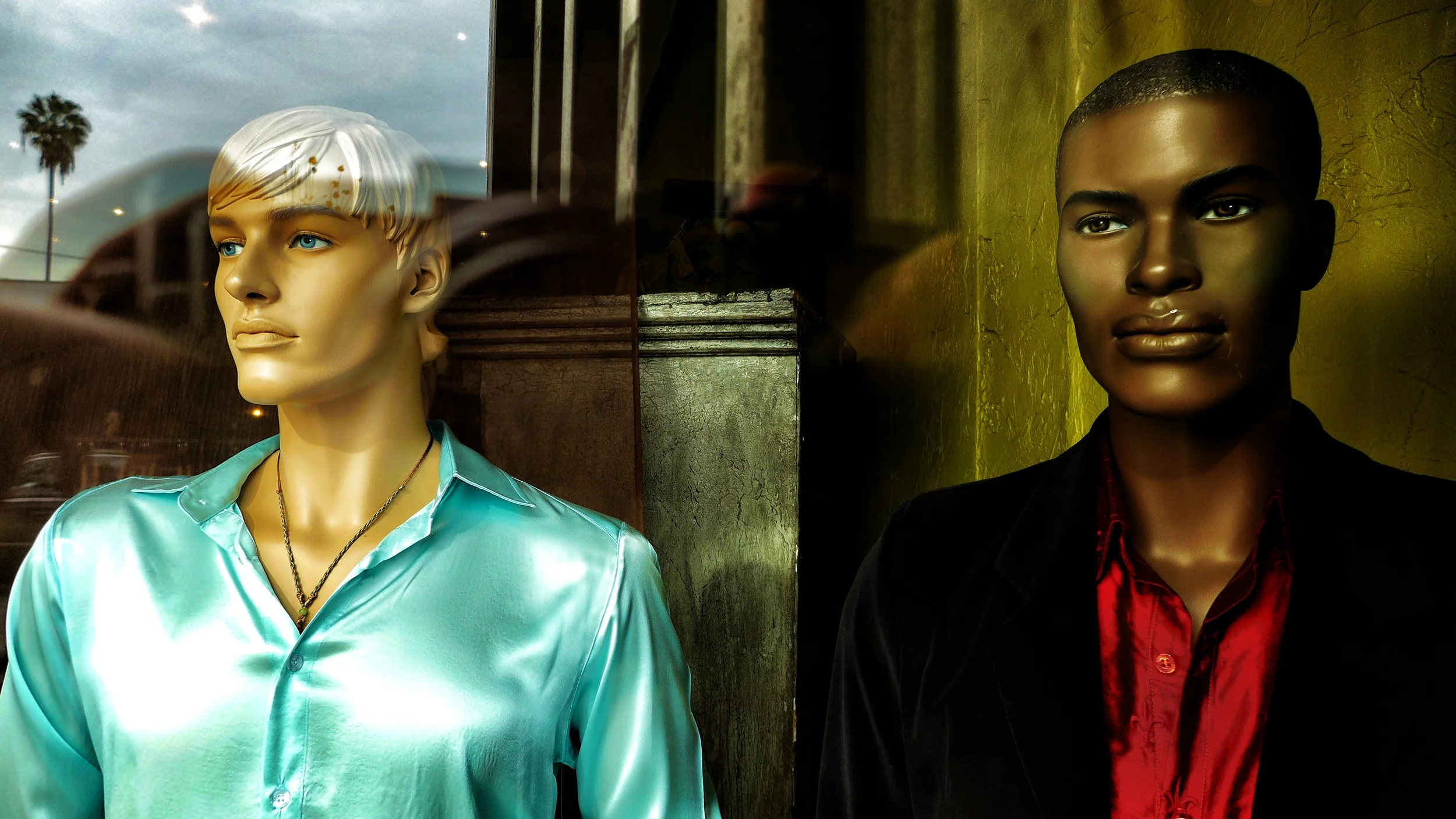 Two mannequins in a store window, one with a blond hairstyle and blue eyes wearing a shiny blue shirt, and the other with short black hair and dark skin wearing a black jacket over a red shirt, with reflections of buildings and palm trees visible in 
