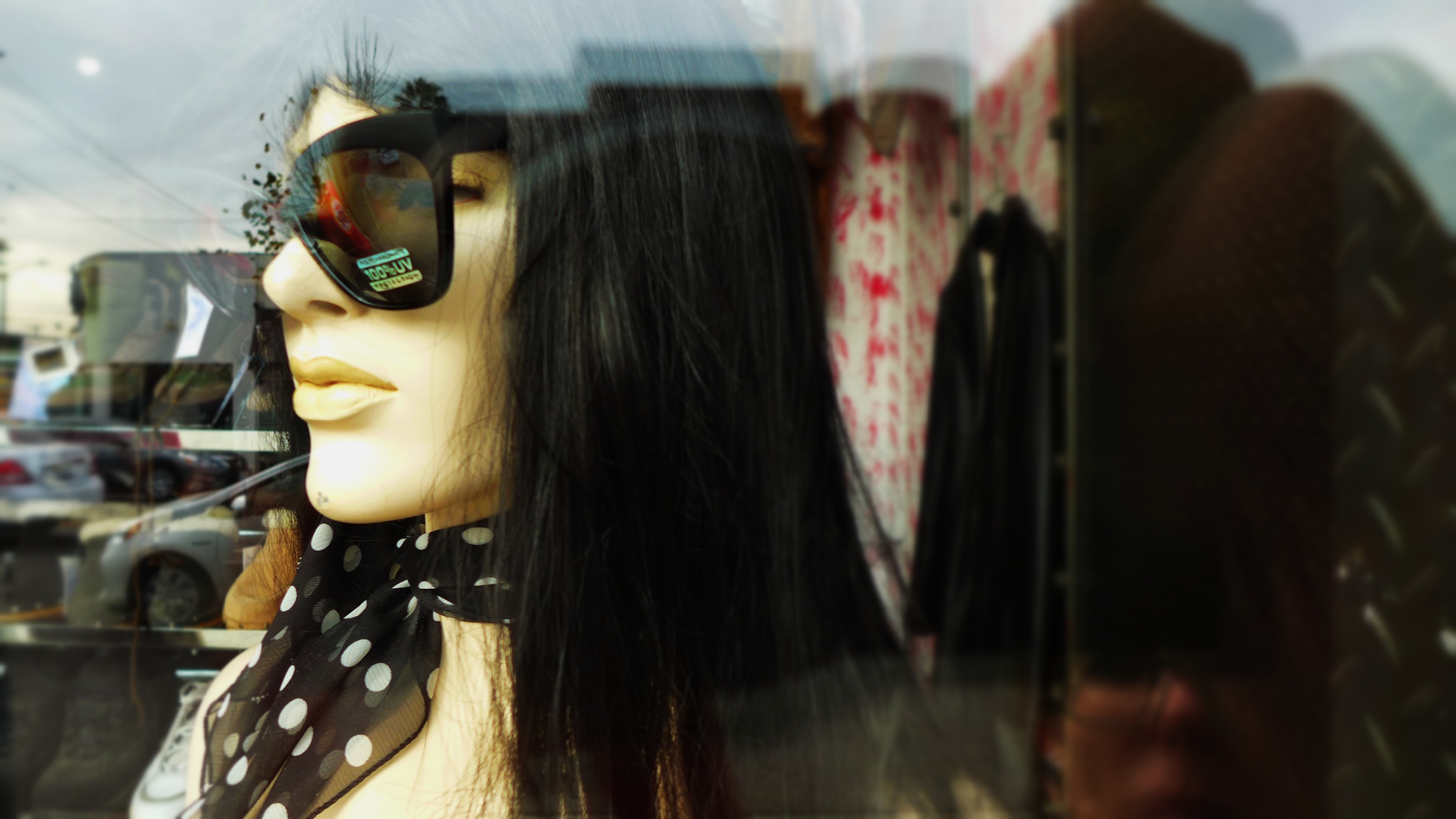A mannequin wearing sunglasses and a polka dot scarf, reflected in a glass display window, with cars and a parking lot outside.
