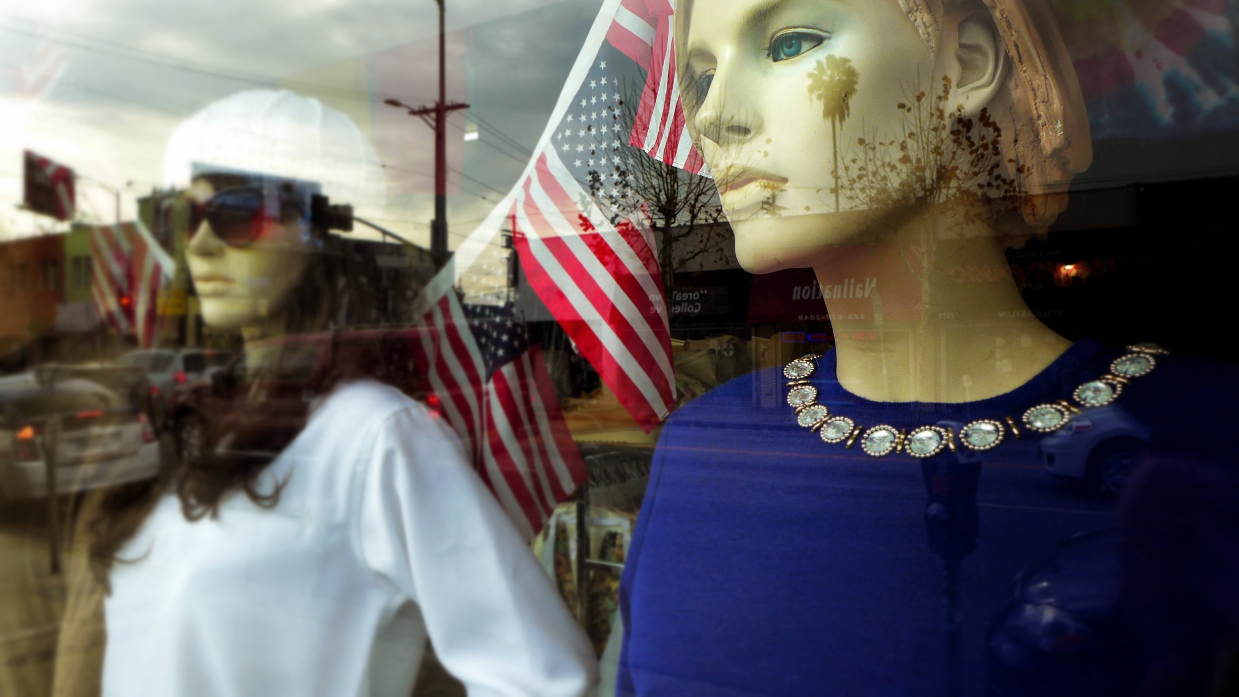 Store window display with a mannequin wearing a blue top and a pearl necklace, and American flags reflected on the glass, along with a partial view of a person with long hair.