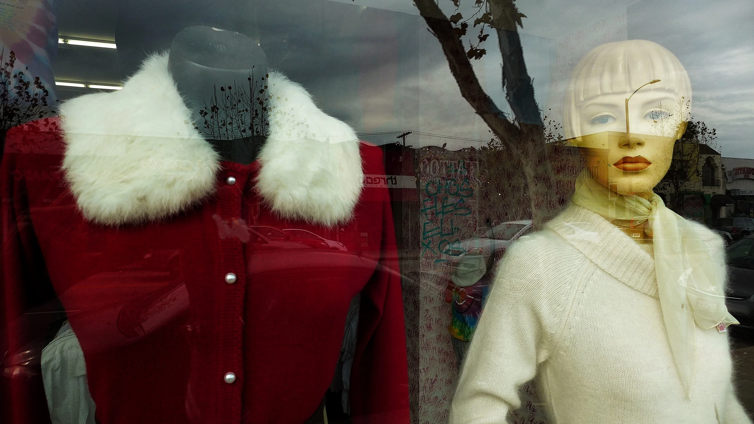Mannequin in store window wearing a red coat with white fur collar and a white sweater with a cream-colored scarf, reflected in the glass with trees, cars, and buildings outside.