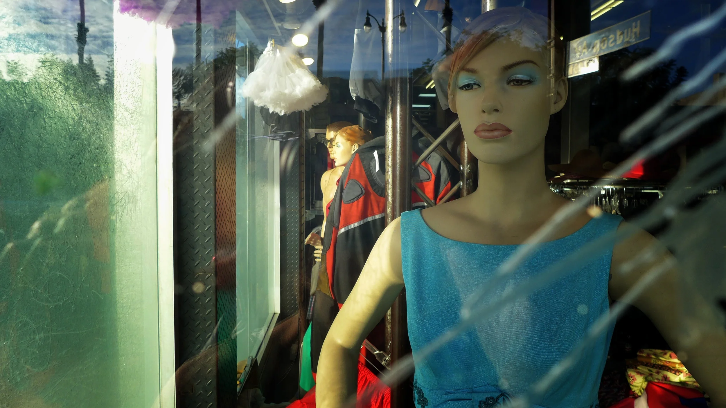 Display window with mannequins dressed in clothing, including a female mannequin in a blue top with makeup and short hair, behind a glass with reflections of trees and sky.