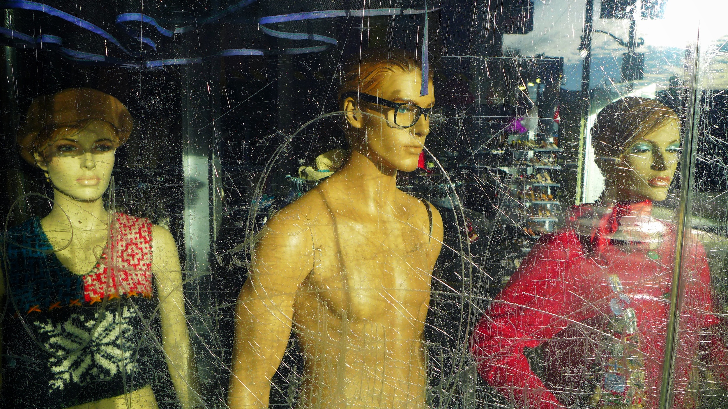 Store window display with mannequins, including one wearing glasses and a black top, seen through a dirty, scratched glass.