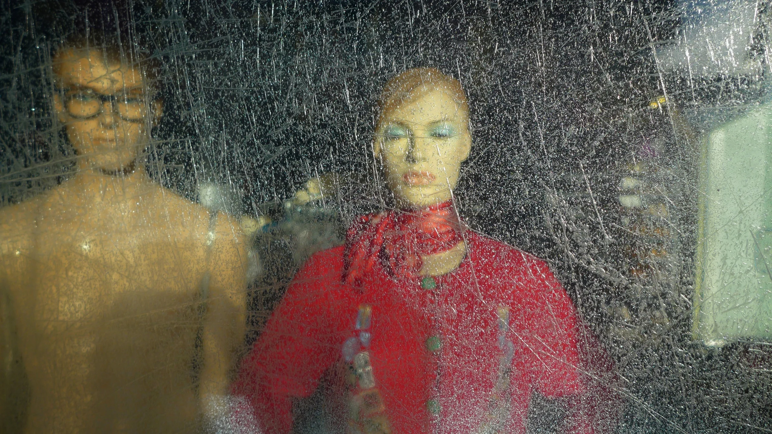 Female mannequin with red hair, red scarf and red dress looking out a scratch-covered window with a bare-chested male mannequin behind her.