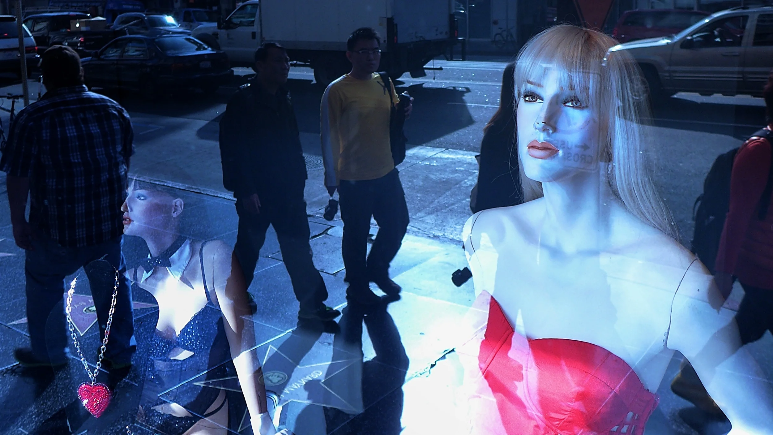 Street scene at night with pedestrians and store window display of mannequins, one with blonde hair wearing a red dress, and young women walking by.