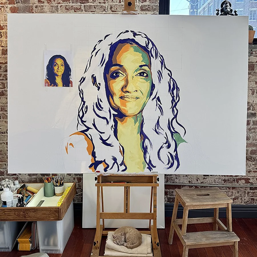 An art studio with a large canvas featuring a colorful, stylized portrait of a woman with curly hair. There is a reference photo of the same woman on the wall, a small wooden stool, a work table with art supplies, and a sleeping cat on a raised platform in front of the canvas.