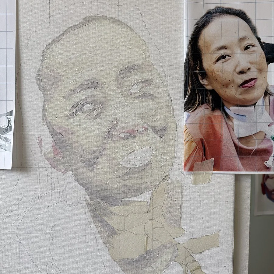 A partially finished painting of a woman’s face on canvas next to a reference photo of the same woman taped to the canvas.
