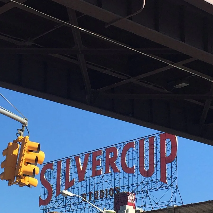 View of a large red sign for Silvercup Studios against a clear blue sky, with a yellow traffic light hanging nearby under an overpass.