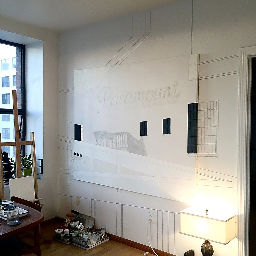 A partially painted wall with the word 'Paramount' faintly visible, some black tiles, and a sketch of a bridge in progress, with a small table lamp and art supplies on the floor.