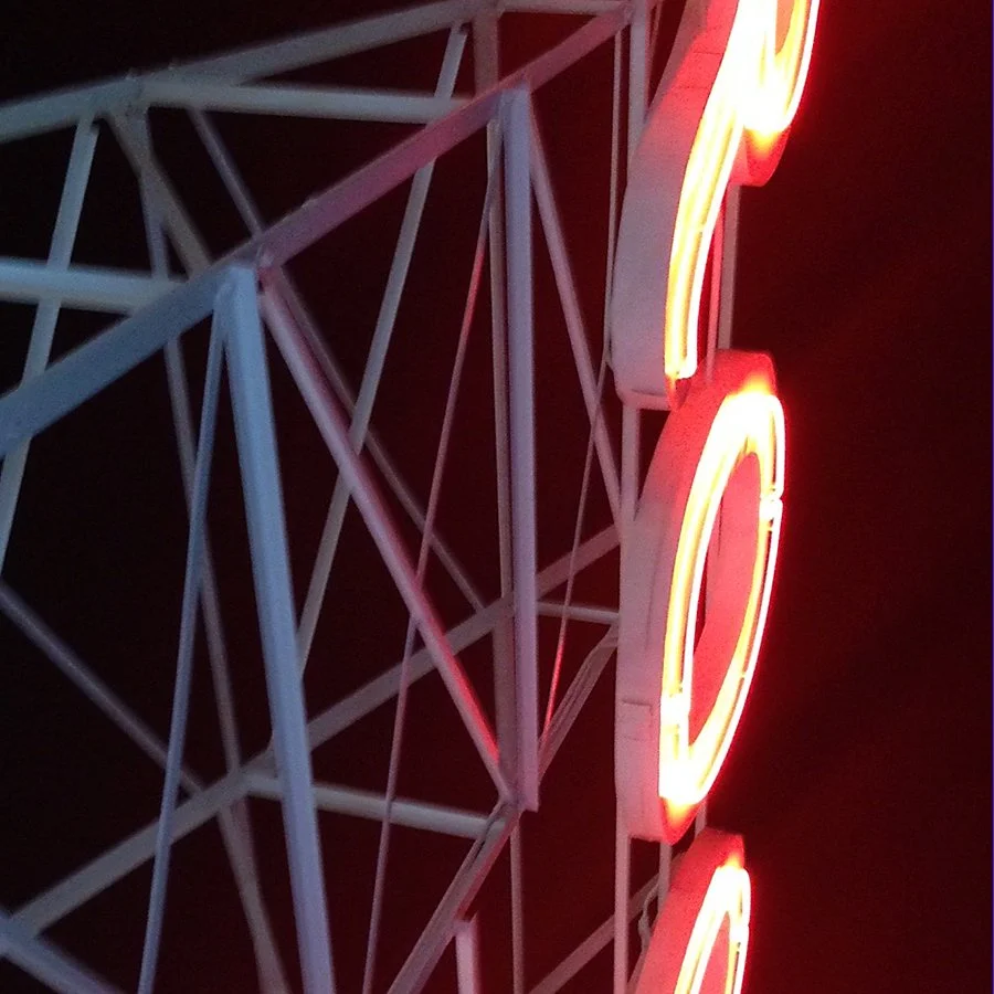 Close-up of a neon sign with bright red and yellow colors, mounted on a white metal structure with intersecting beams against a dark night sky.