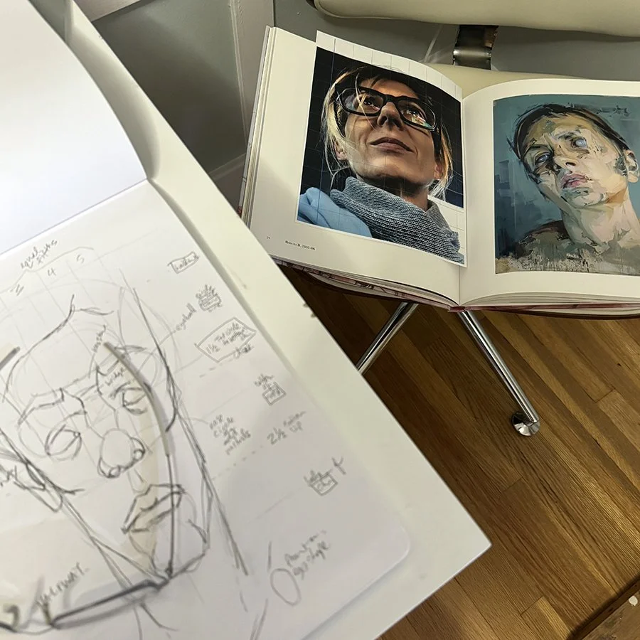 Open book with portrait photos of women, one wearing glasses and scarf, the other with short hair and makeup, on a wooden surface with a sketchpad nearby.