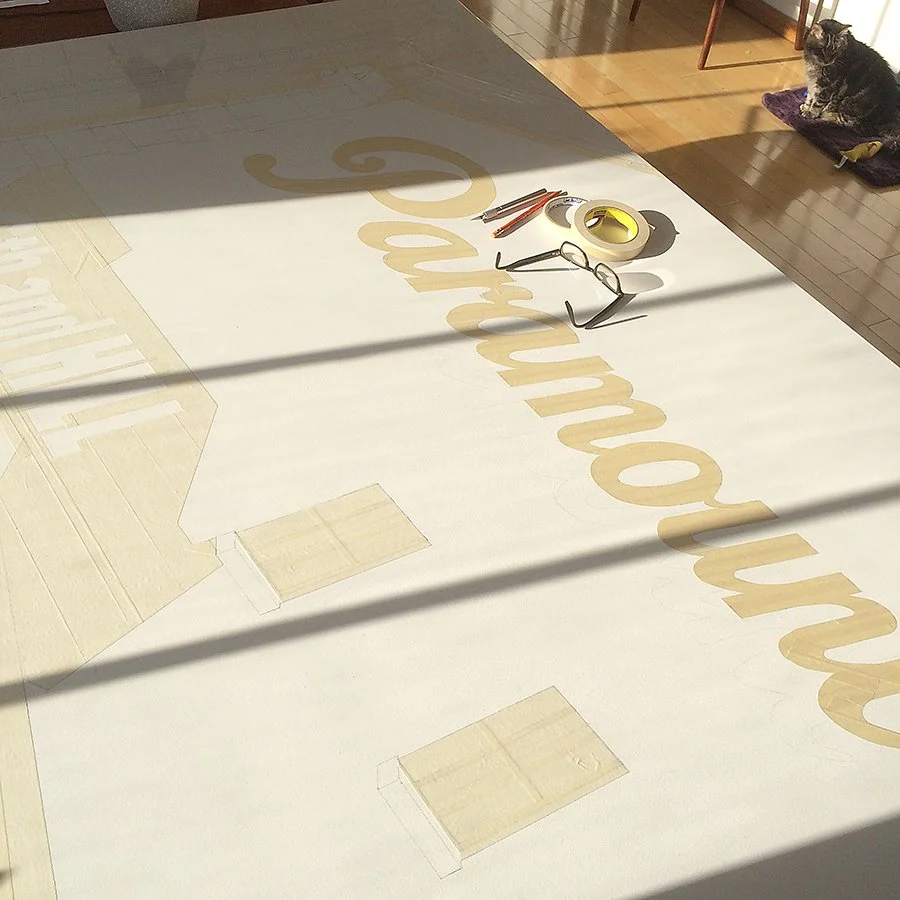 Large white poster paper with the partially visible word "Welcome" in golden letters, placed on a wooden floor. Items on the paper include a pen, a pair of glasses, and a tape dispenser. In the background, a cat is sitting on a purple cushion near a wooden chair.
