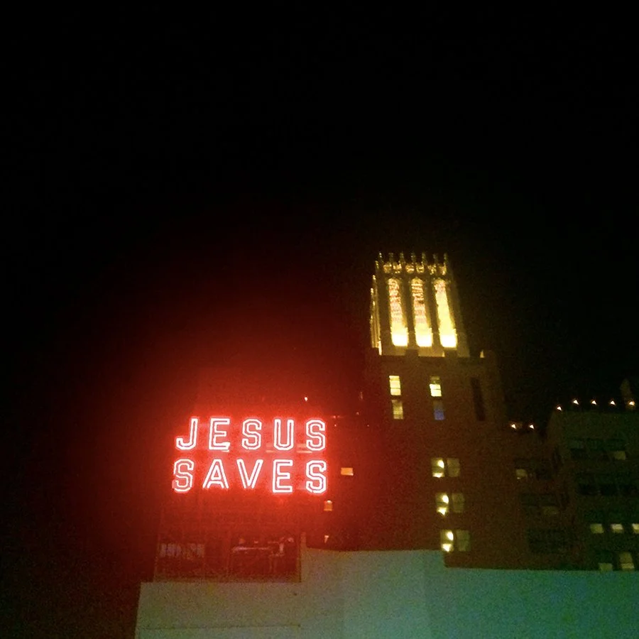 Nighttime city scene with a bright red neon sign that says 'Jesus Saves' and a tall illuminated tower in the background.