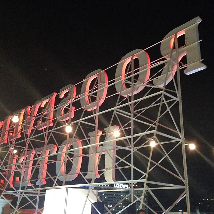 Nighttime view of a large, illuminated sign marked "Salvador" with a metal framework, string lights, and a city skyline in the background