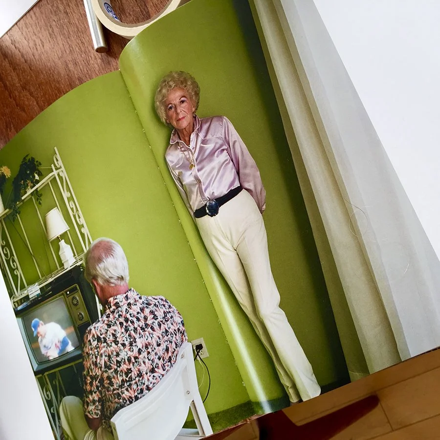 A woman wearing a pink satin blouse and cream pants standing beside a green wall, with an elderly man with white hair watching television in the foreground.