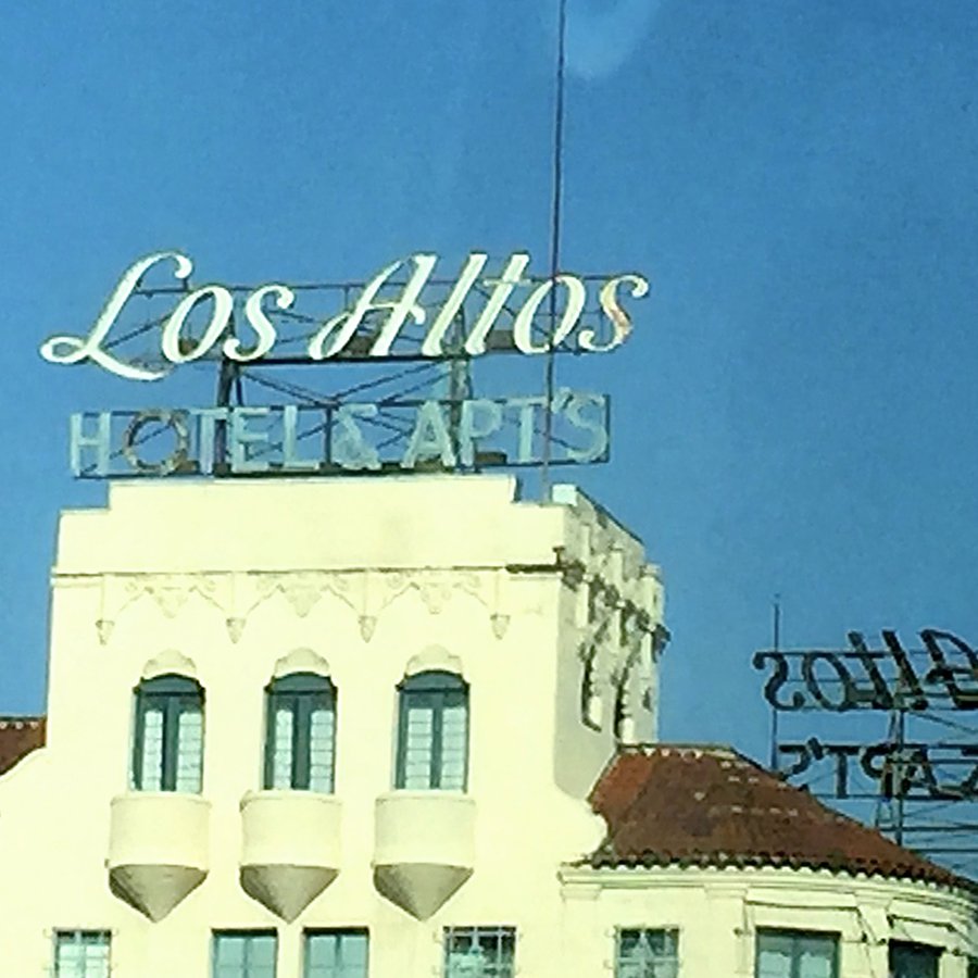 The image shows the top of a white building with three arched windows, with a neon sign on top that reads 'Los Alta's HOTEL APARTS' against a blue sky.