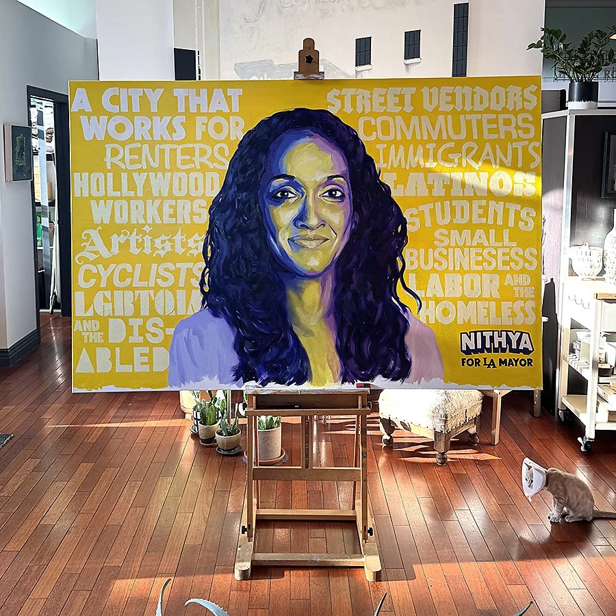 A colorful mural of a woman with dark, curly hair, set against a yellow background with text supporting Nithya for LA mayor. The mural is indoors on a wooden floor with plants and furniture visible around.