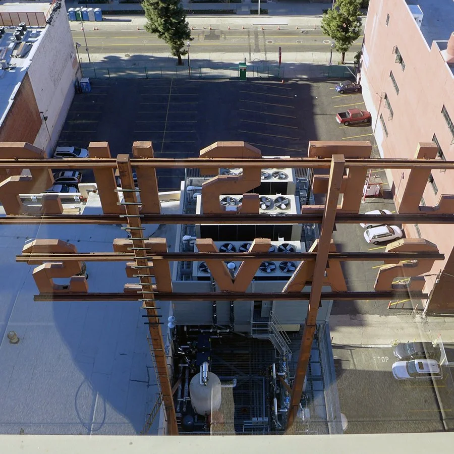 View from a high-rise building overlooking a parking lot and street. The rooftop HVAC unit with large fans is visible in the foreground, with the word 'LOVE' in large orange letters above it. Two trees are also seen in the parking lot.