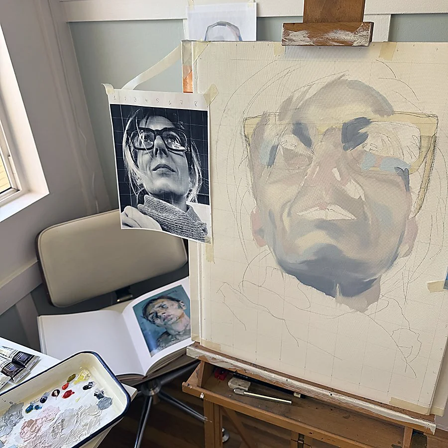 An artist's workspace with a partially painted portrait of a person wearing glasses, a printed reference photo of the same person taped to the canvas, a book with another portrait photo, and a palette with paints.