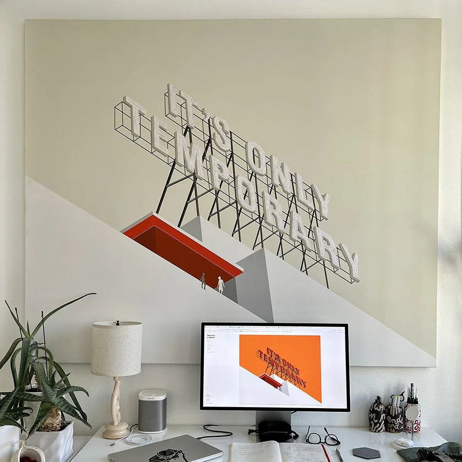 A large wall art piece featuring a geometric design with stairs and bold text reading 'This is a very interesting project' in 3D letters, situated above a white desk with a computer, lamp, and various desk items.
