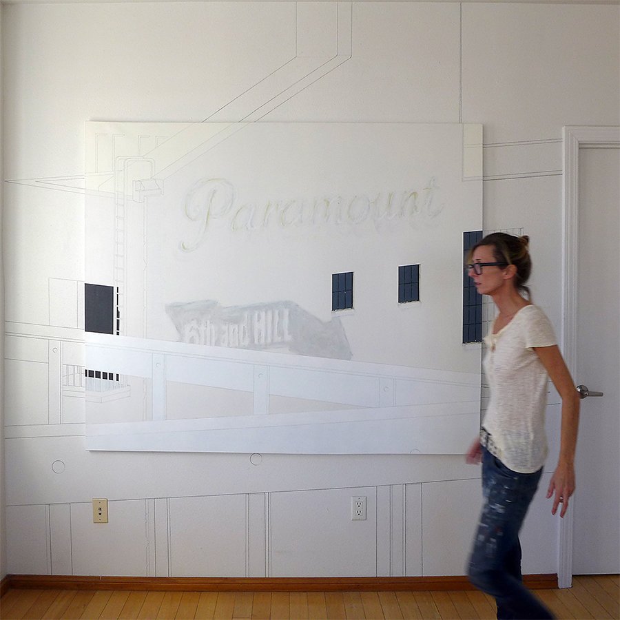 A woman walking past a wall with a large white canvas or paper covering part of a schematic or technical drawing of a building, including windows, doors, and plumbing or electrical lines. The scene appears to be in an office or art studio.