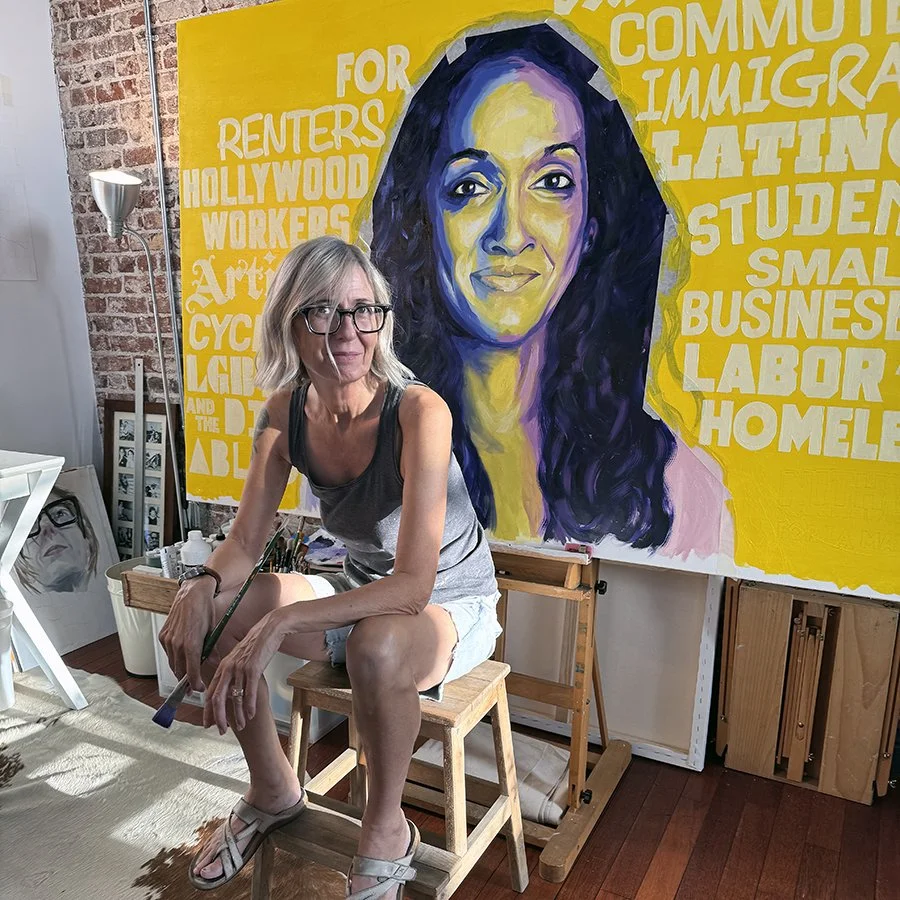 A woman with gray hair, glasses, and a casual outfit is sitting on a wooden stool in front of a colorful wall painting of a woman's face. The background includes words related to community and immigration.