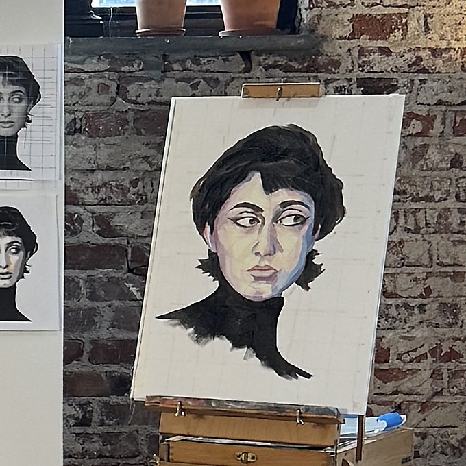 An art studio with a portrait of a woman on a canvas, showing a painted interpretation of a reference photo pinned on a white board. Two potted cacti sit on the window sill, and a brick wall is visible in the background.