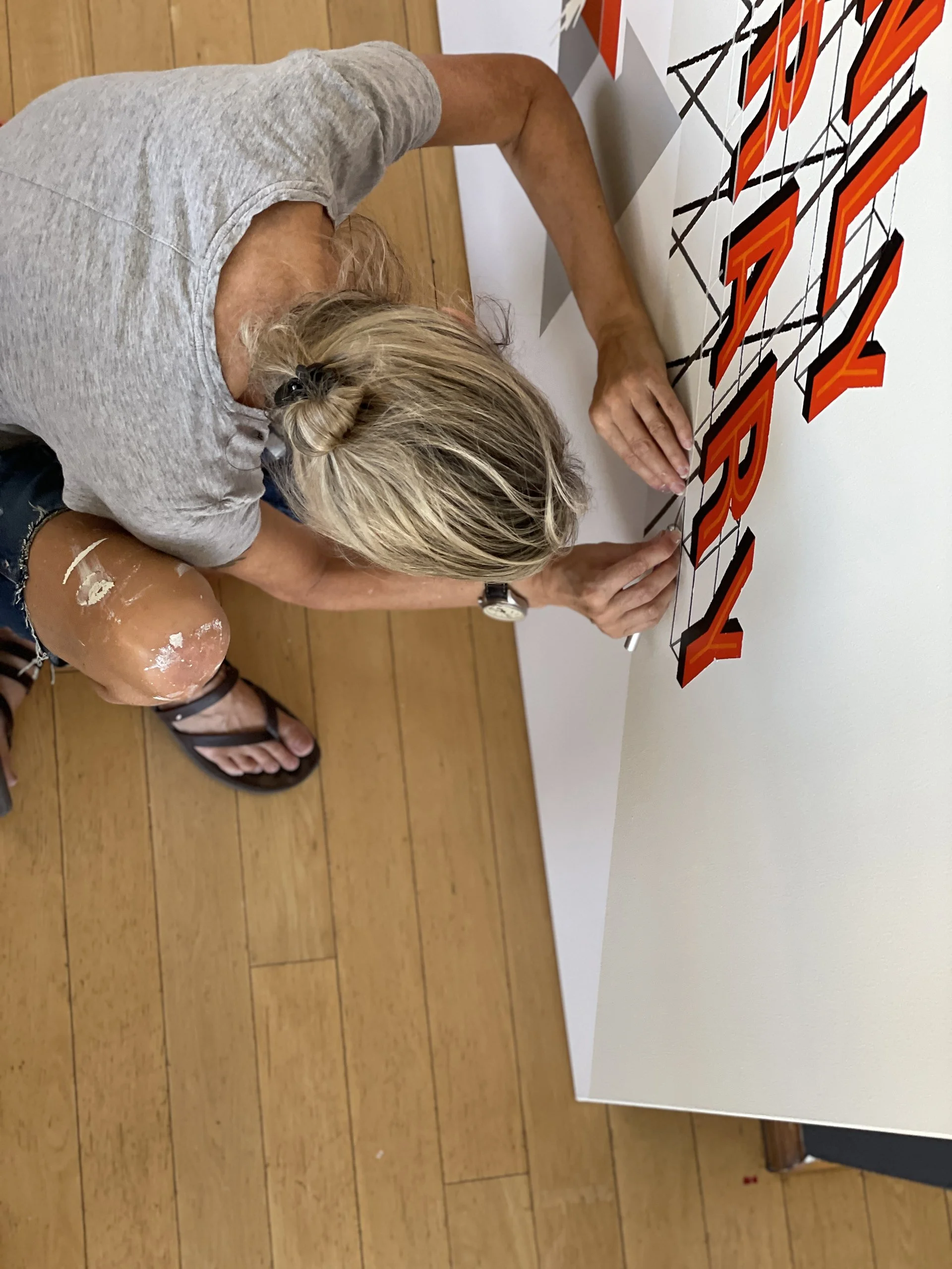 The artist, Daisy Tong, photographed from above, with blonde hair, wearing a gray t-shirt, ripped jeans, and sandals, sitting on the wooden floor, working on a large art piece on a white wall with orange, black, and white patterns.