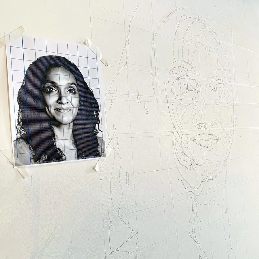 A pencil sketch of a woman’s face on a wall with a reference photo taped next to it.
