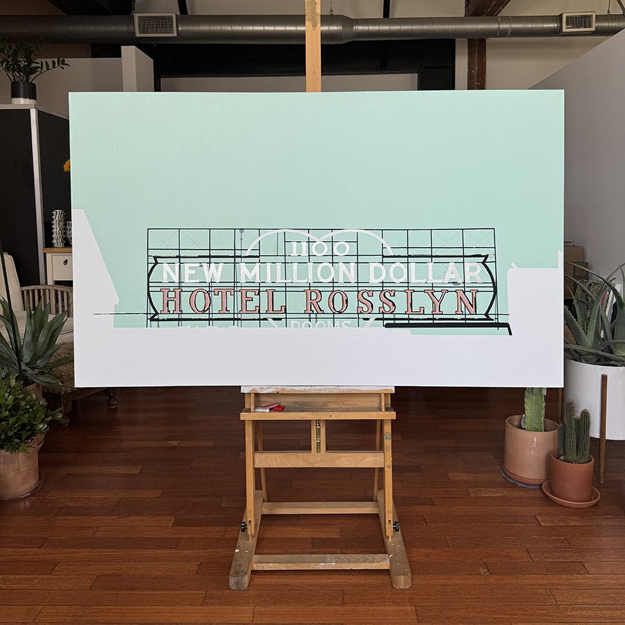 Easel with a sign that reads "New Million Dollar Hotel Rosslyn" in front of a white wall with plants on each side.