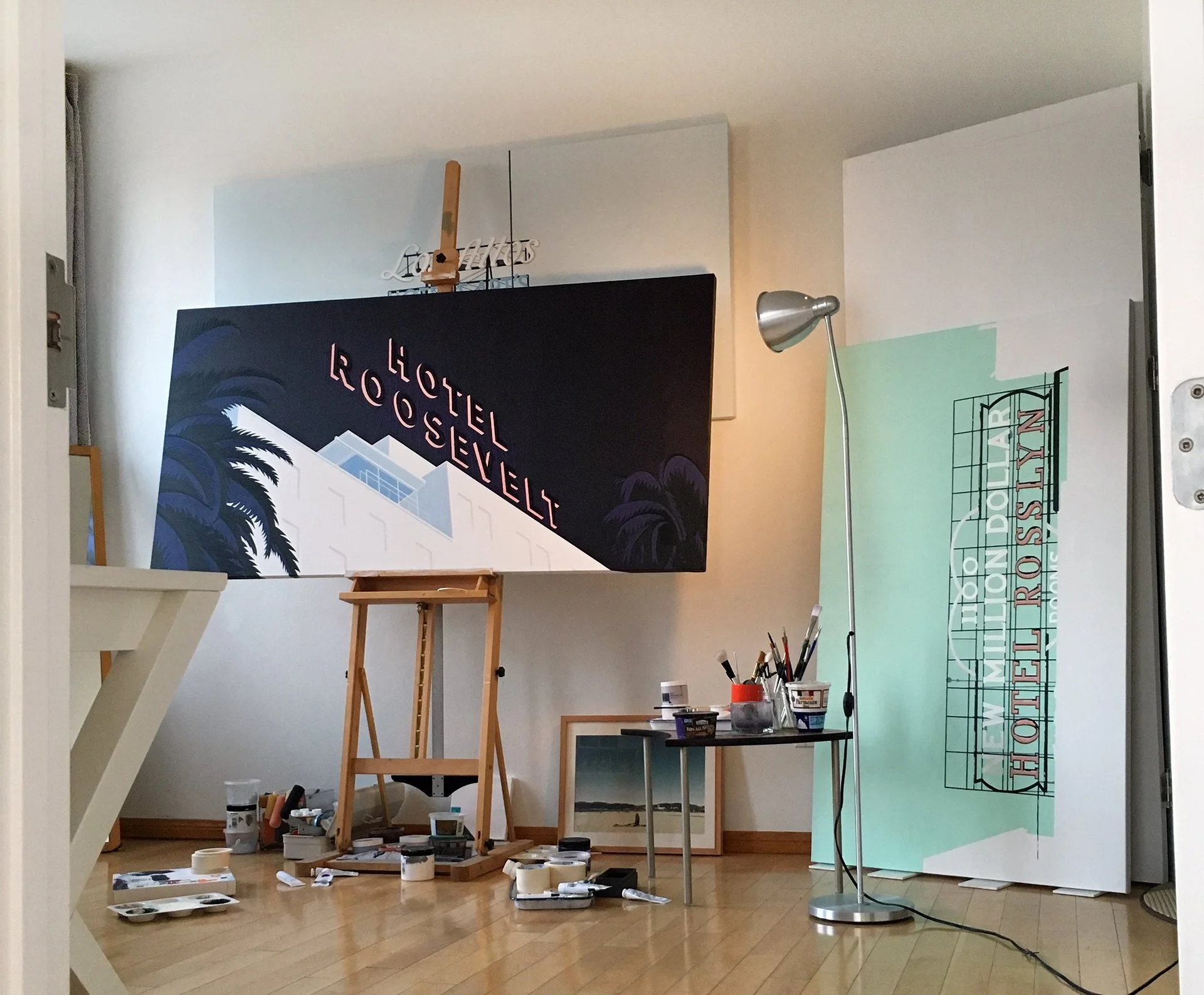 An art studio with a painting on an easel and paint supplies on the floor. The space has hardwood floors and a white wall with a lamp.