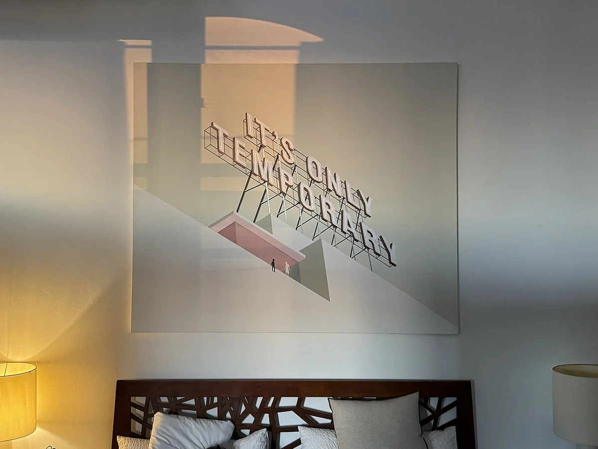A framed artwork hangs on a white wall above a bed with pillows, with two small lamps on either side, one lit with warm yellow light.