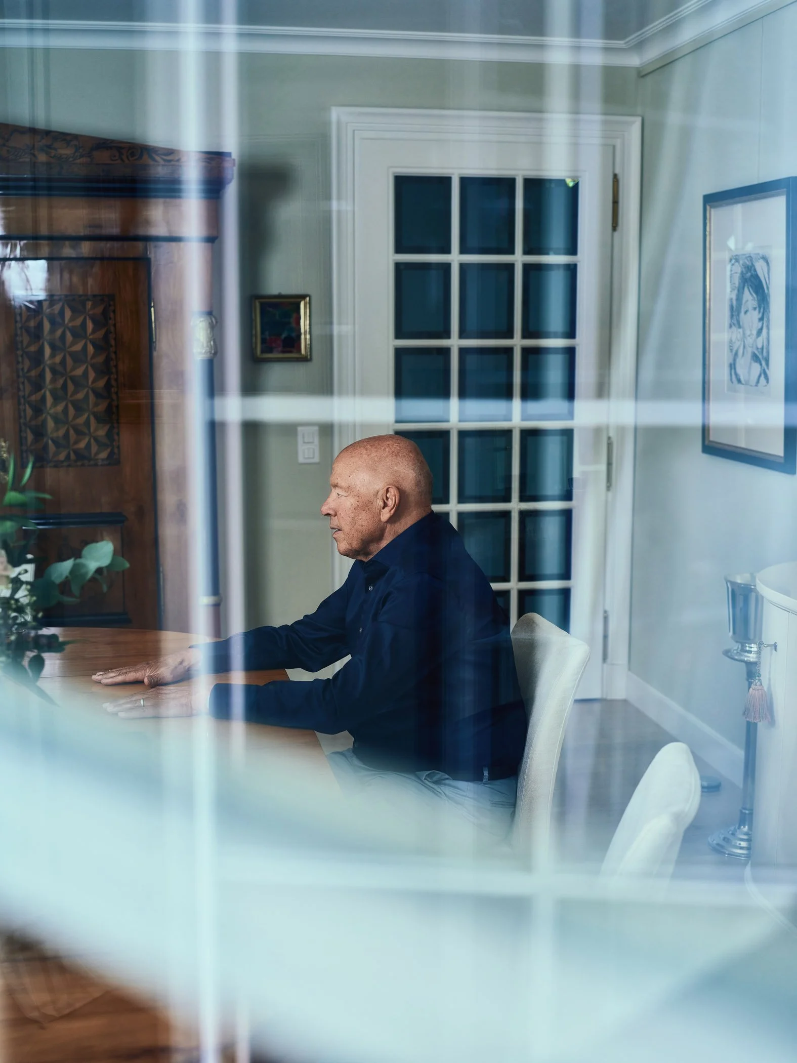 Theo Müller im Profil an einem Tisch, fotografiert durch eine spiegelnde Fensterscheibe.