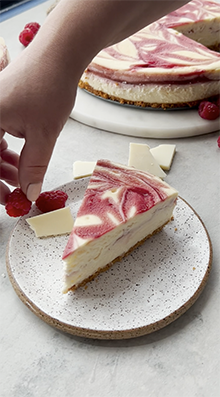 a hand places a raspberry next to a slice of cheesecake