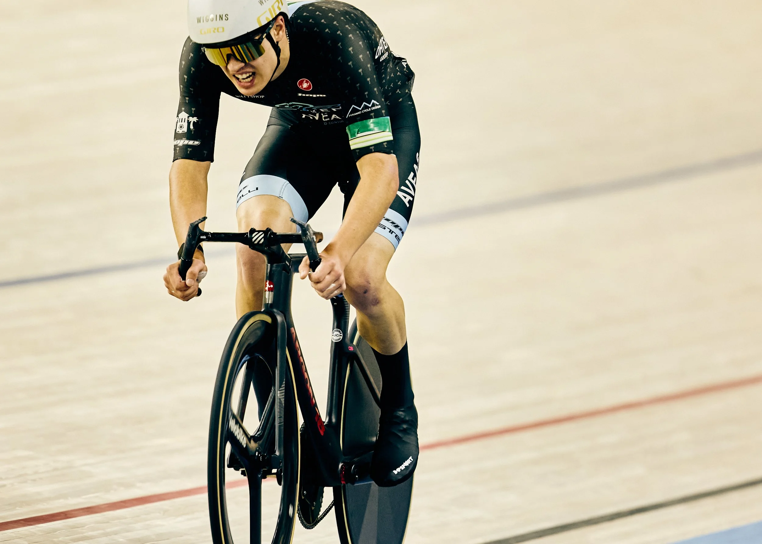 A male cyclist races on an indoor velodrome track, wearing a black and white racing suit, a white helmet, and sunglasses, focused and exerting effort.