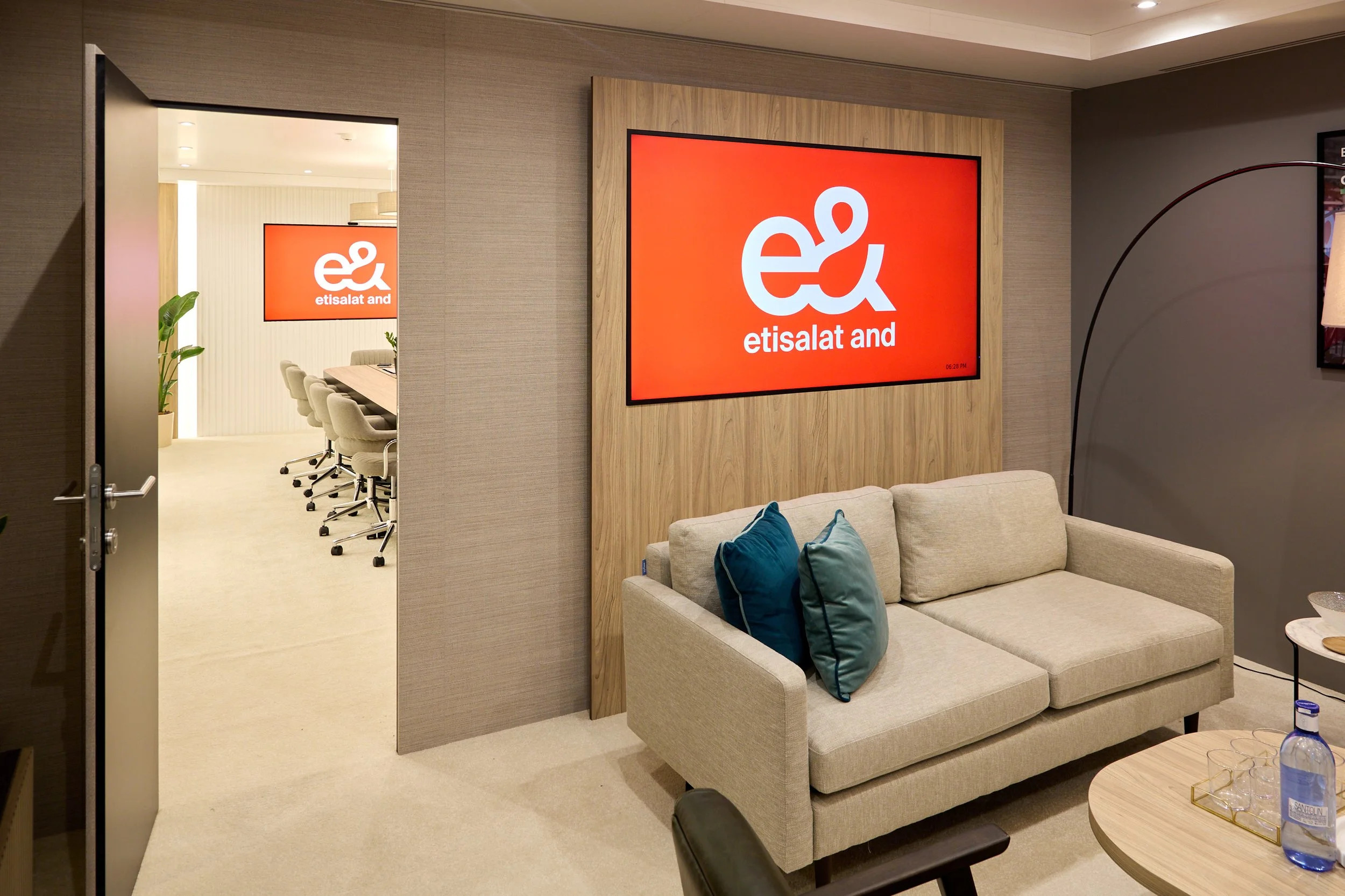 A modern hotel lounge with a beige sofa, blue and teal pillows, a wooden coffee table with water bottles, a large wall-mounted screen displaying the 'etisalat and' logo, and an open door leading to a conference room with chairs and another screen.