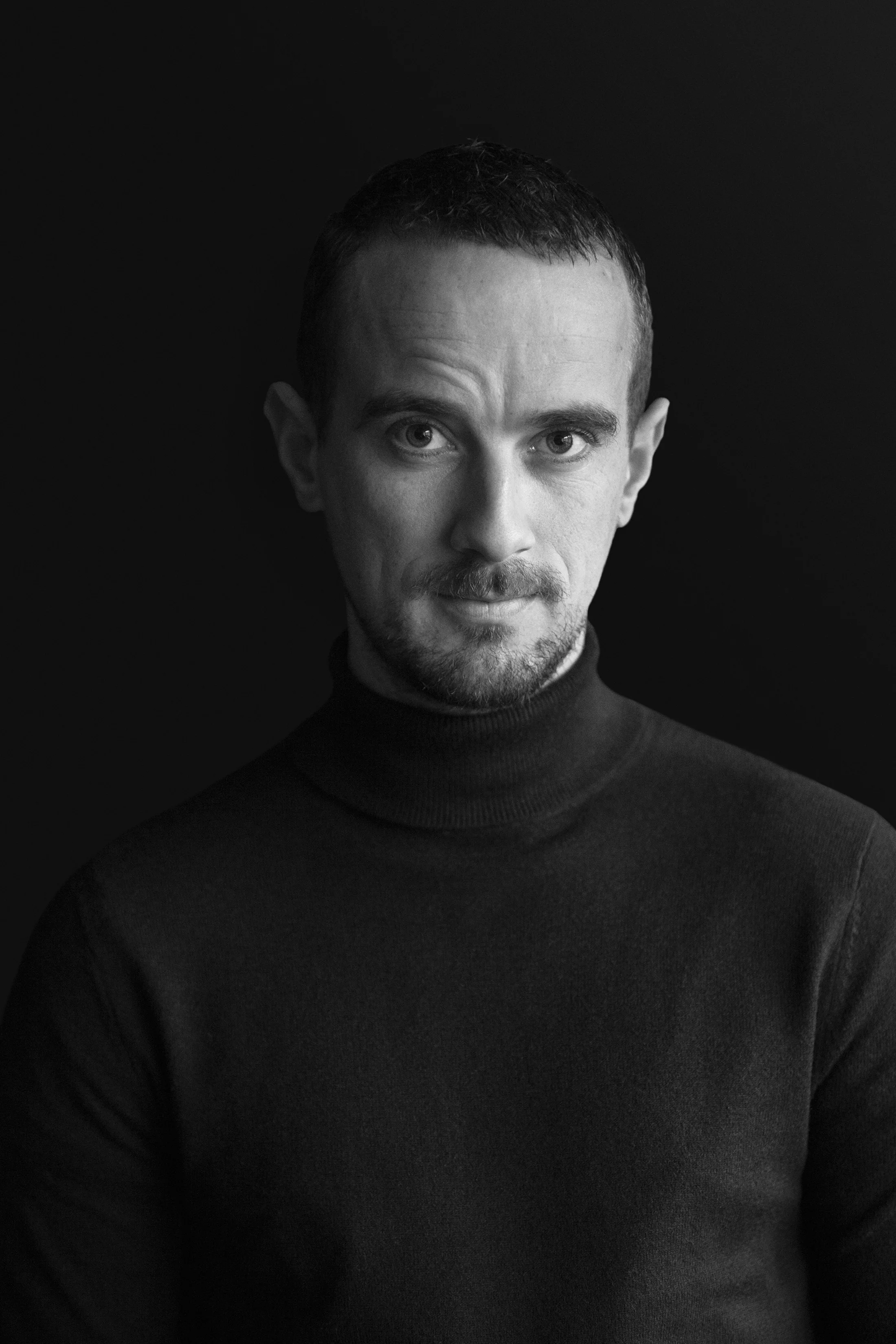 Black and white portrait of a man with short hair, wearing a dark turtleneck sweater, against a dark background.