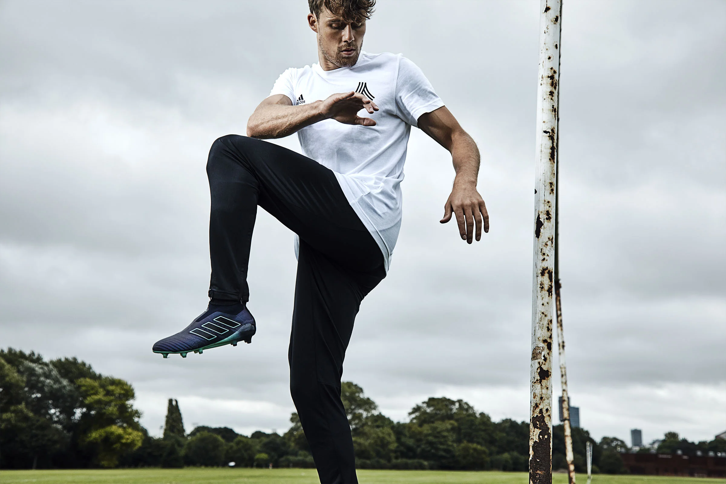 A young man in athletic wear stretching his leg on a soccer field, standing next to a rusty goalpost with overcast sky and trees in the background.