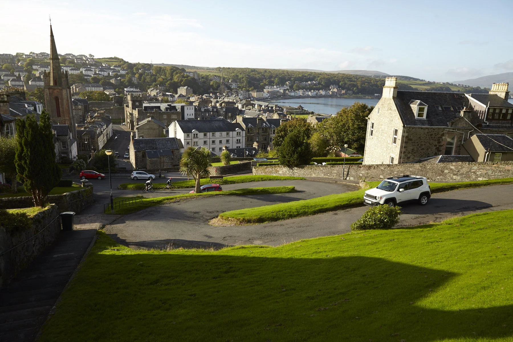 A scenic view of a coastal town with a hill, a church steeple, houses, a bay, and greenery, including a well-kept lawn, a white SUV, and a curved road with a few motorcycles and cars.