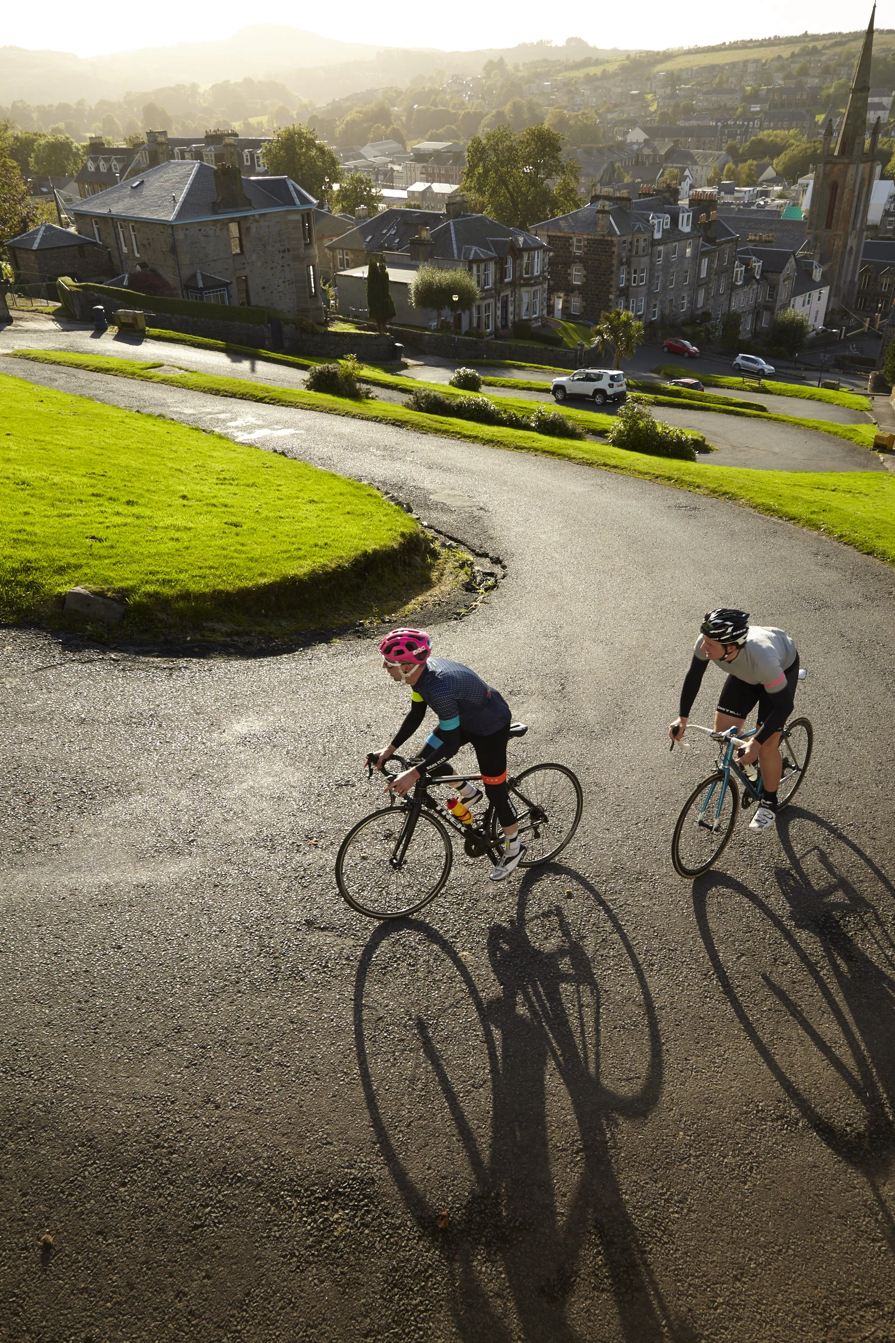 Two cyclists riding on a winding road through a scenic, hilly, suburban area with greenery and houses in the background, during a sunny day.