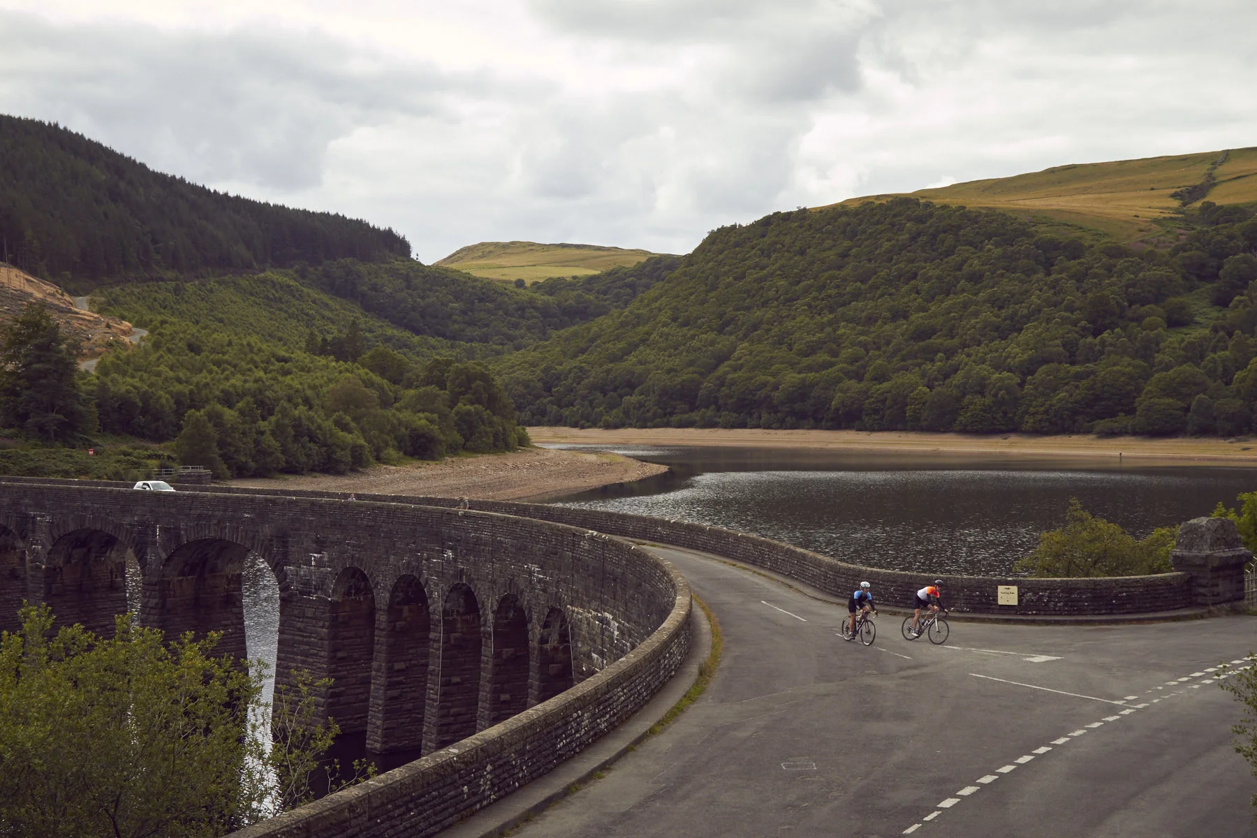 Two cyclists riding on a curved road next to a stone bridge over a reservoir, surrounded by green hills and a cloudy sky.