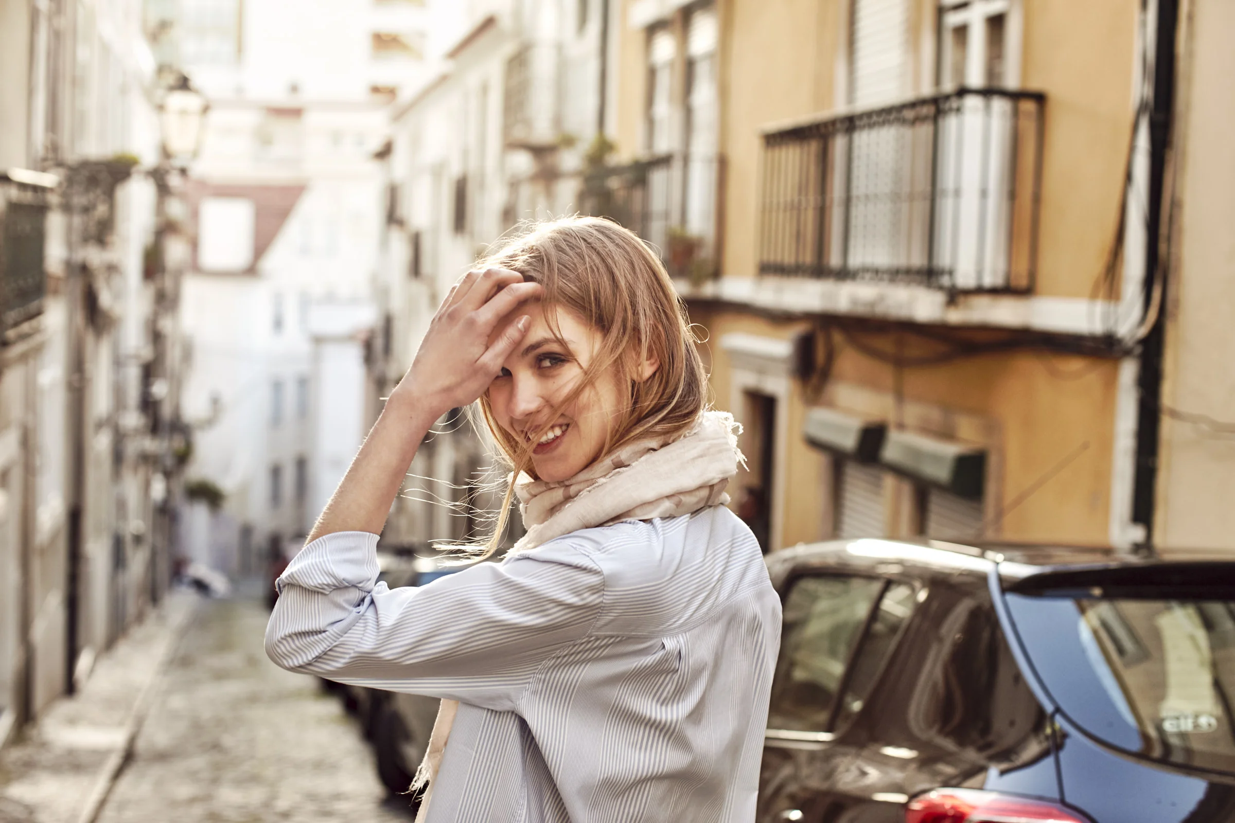 A smiling young woman with blonde hair and a beige scarf around her neck, standing on a cobblestone street in an urban area with European-style buildings and parked cars, during daylight.