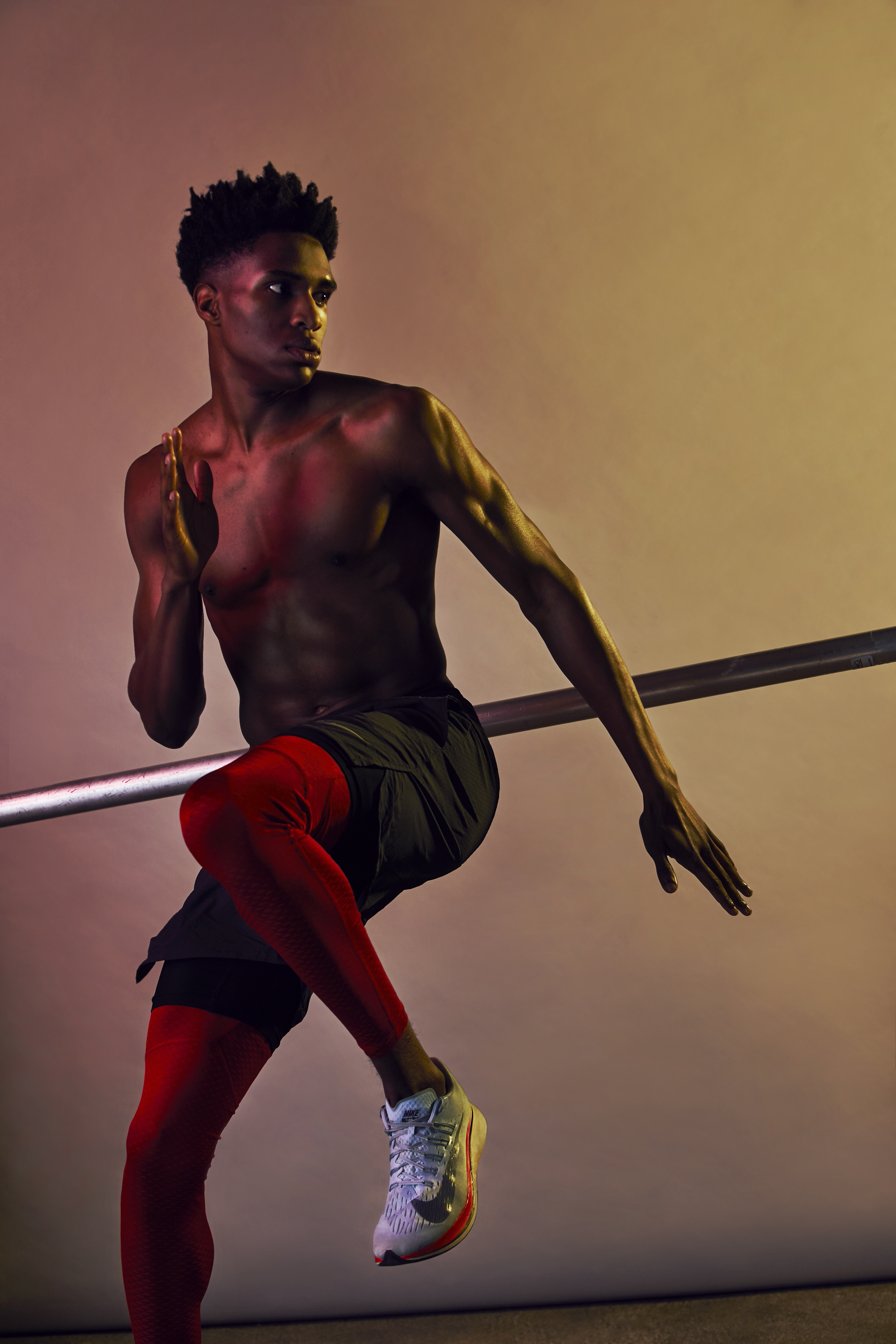 A shirtless young man in athletic shorts and running shoes balancing on a bar with one leg raised, against a plain gradient background.