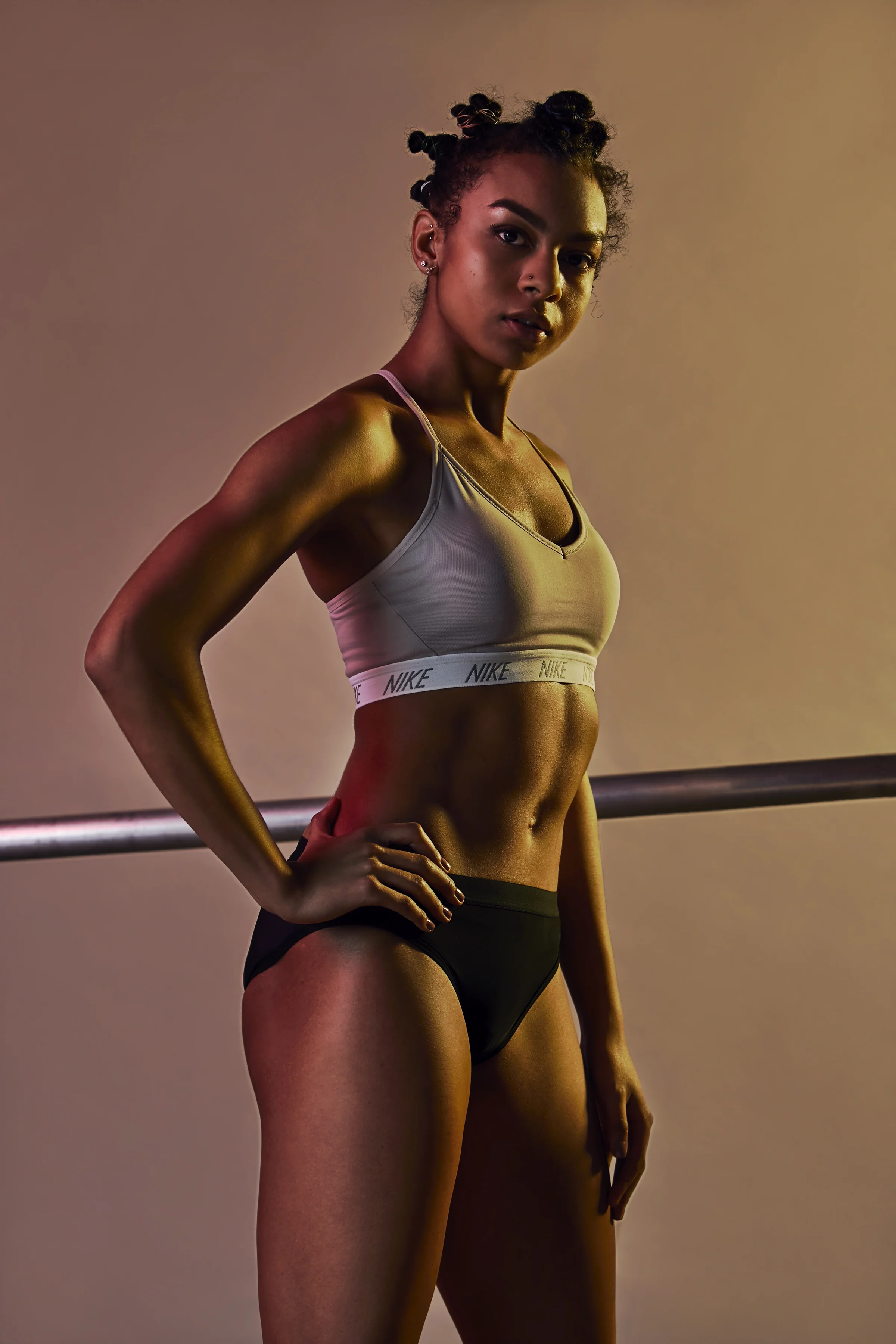 A fit woman with dark skin and curly hair styled in bantu knots standing next to a ballet barre, wearing a beige Nike sports bra and black underwear, looking at the camera.