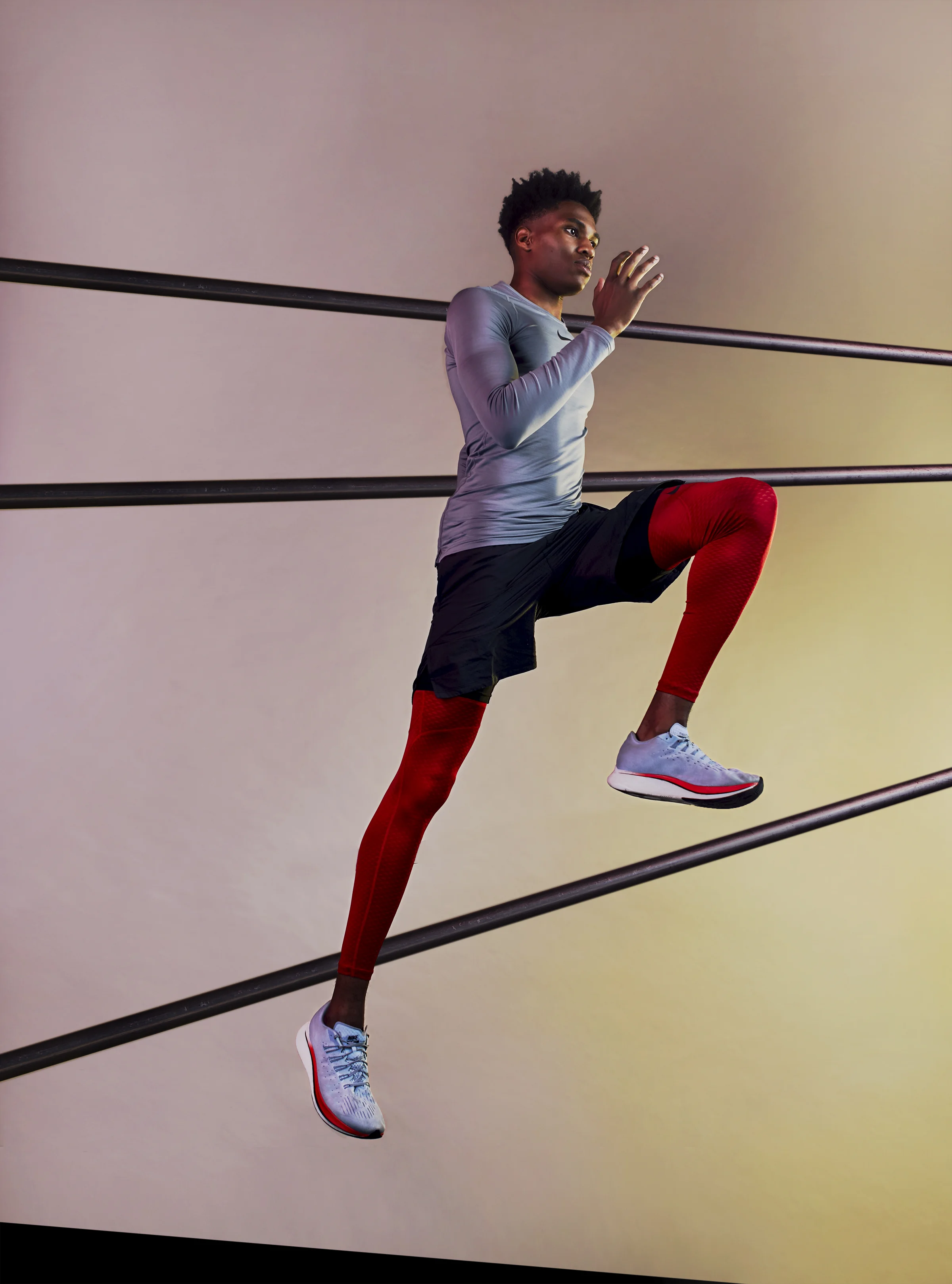 An athlete in a gray long-sleeve shirt and black shorts performing a high knee exercise on a horizontal bar, wearing red compression leggings and white running shoes, with a neutral background.
