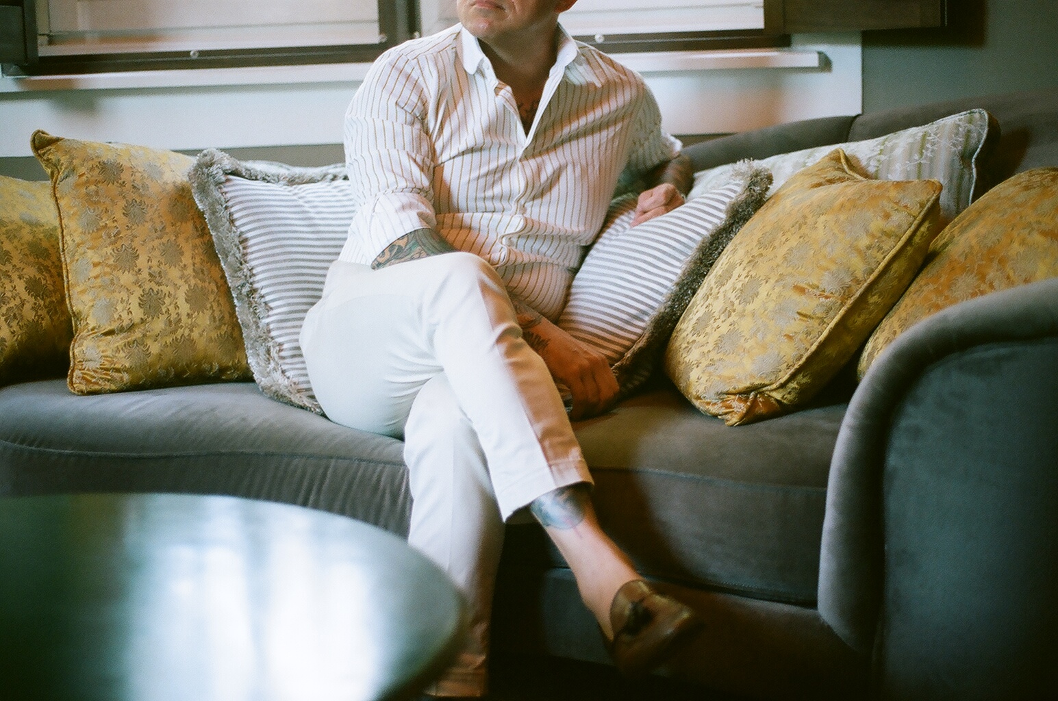A man sitting on a couch with multiple decorative pillows, wearing a striped shirt and white pants, with tattoos visible on his arm and leg, in a modern living room.