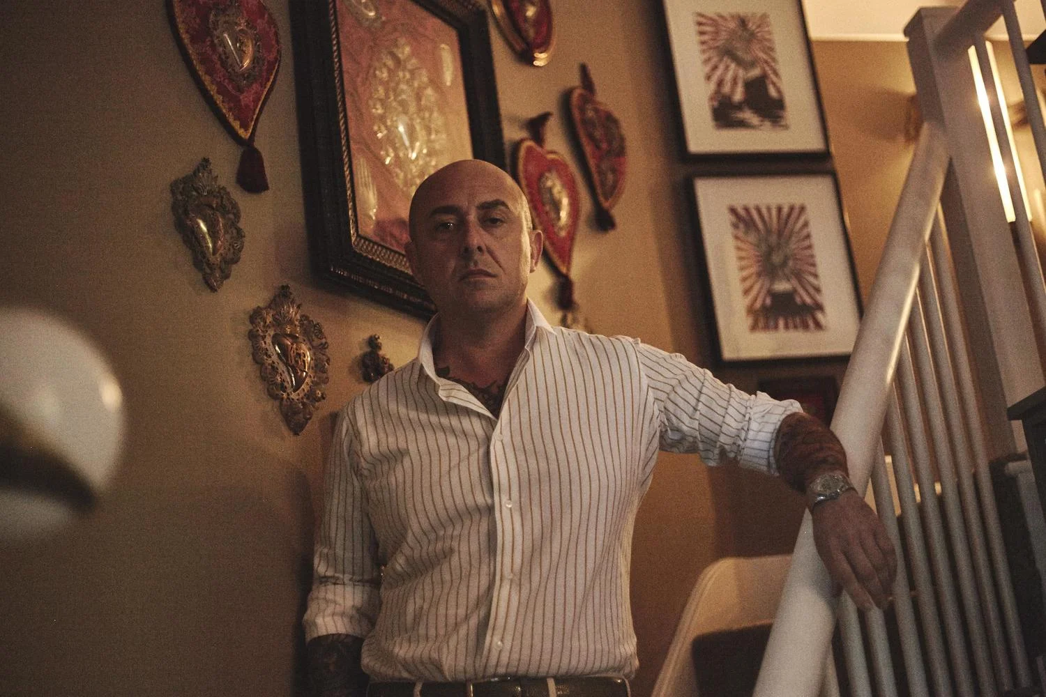 A bald man wearing a striped white shirt and a watch leans against a staircase railing in a room decorated with framed artwork and heart-shaped wall decorations.