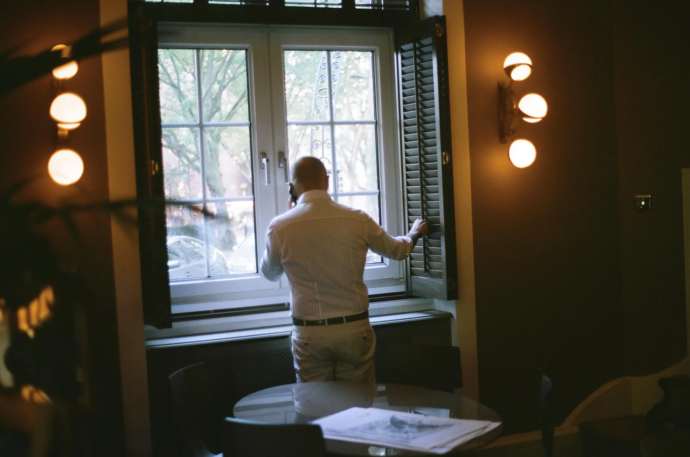 A man stands at a window, looking outside and holding the window shutter open, in a warmly lit room with walls illuminated by a wall sconce with four round lights.