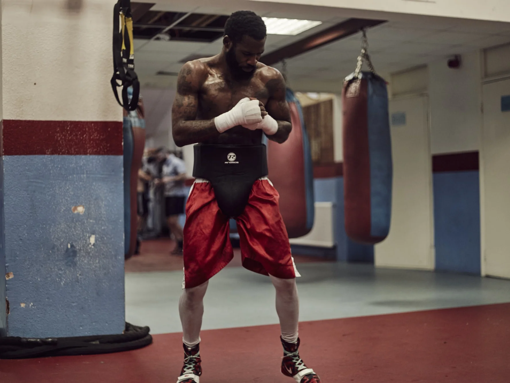A shirtless male boxer with tattoos stands in a gym, wearing red shorts, boxing gloves, and leg wraps, preparing for training near punching bags.