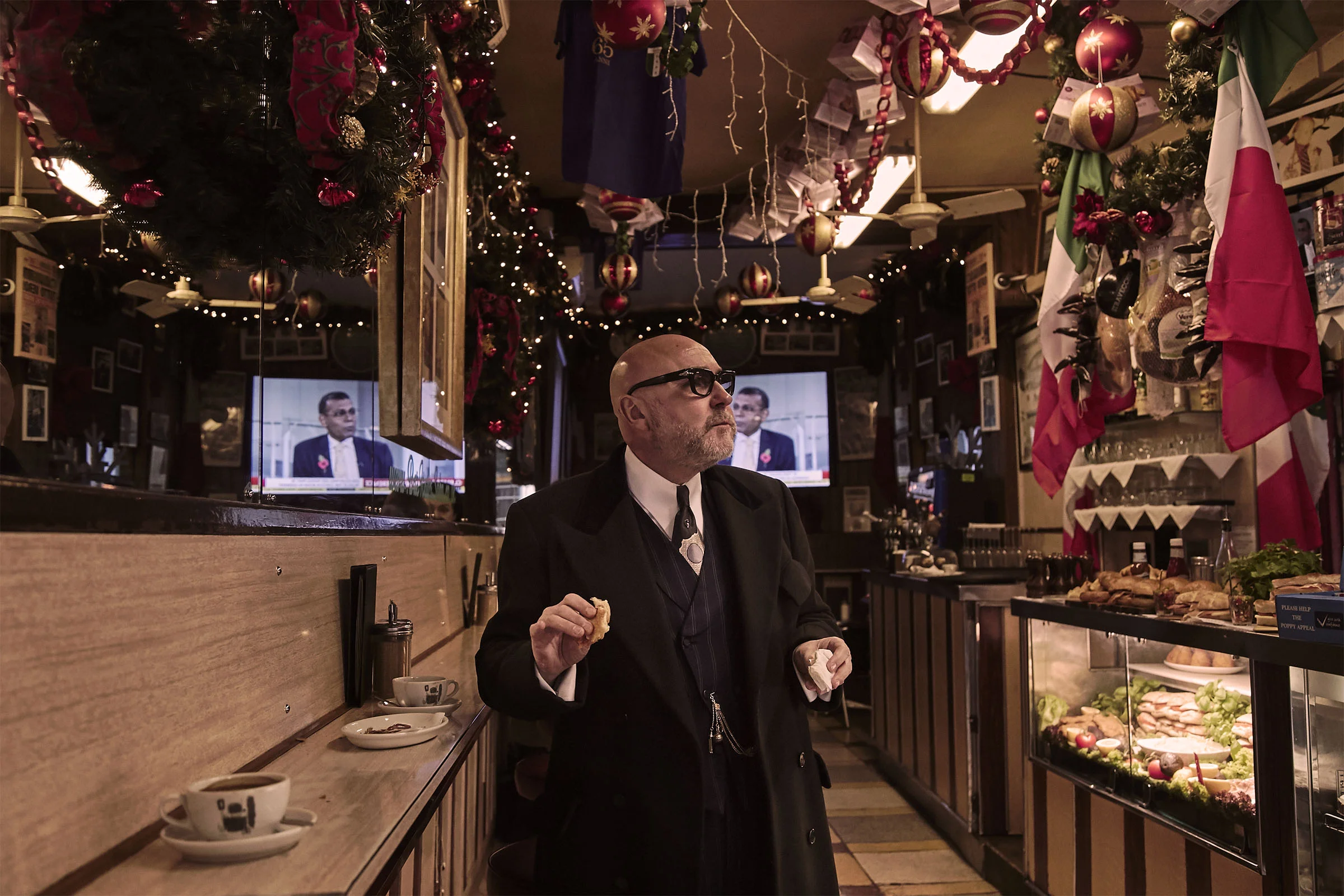 A man in a black suit, glasses, and white shirt eating in a bar decorated for Christmas with ornaments, flags, and lights, with two TV screens in the background.